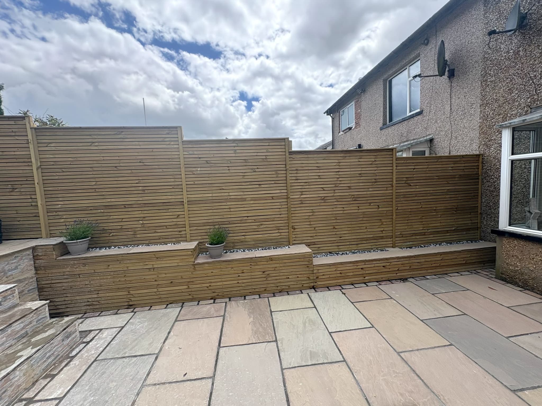 Stepped slatted timber fence above raised timber planters and a sandstone patio